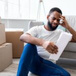 Frustrated African Man Reading Bill Sitting Among Moving Boxes Indoor