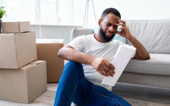 Frustrated African Man Reading Bill Sitting Among Moving Boxes Indoor